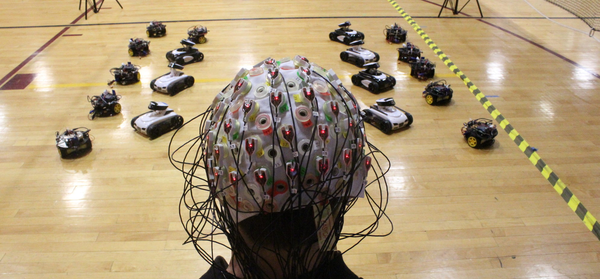 Wheeled robots on a gym floor in front of a person wearing a cap with wires and electrodes on it